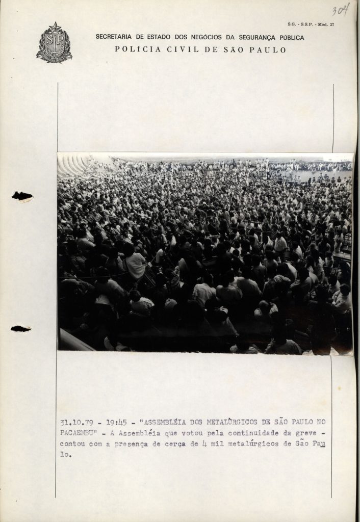: Foto em preto e branco uma uma arquibancada e parte do campo de um estádio de futebol lotado de pessoas. Todos olham para a mesma direção.