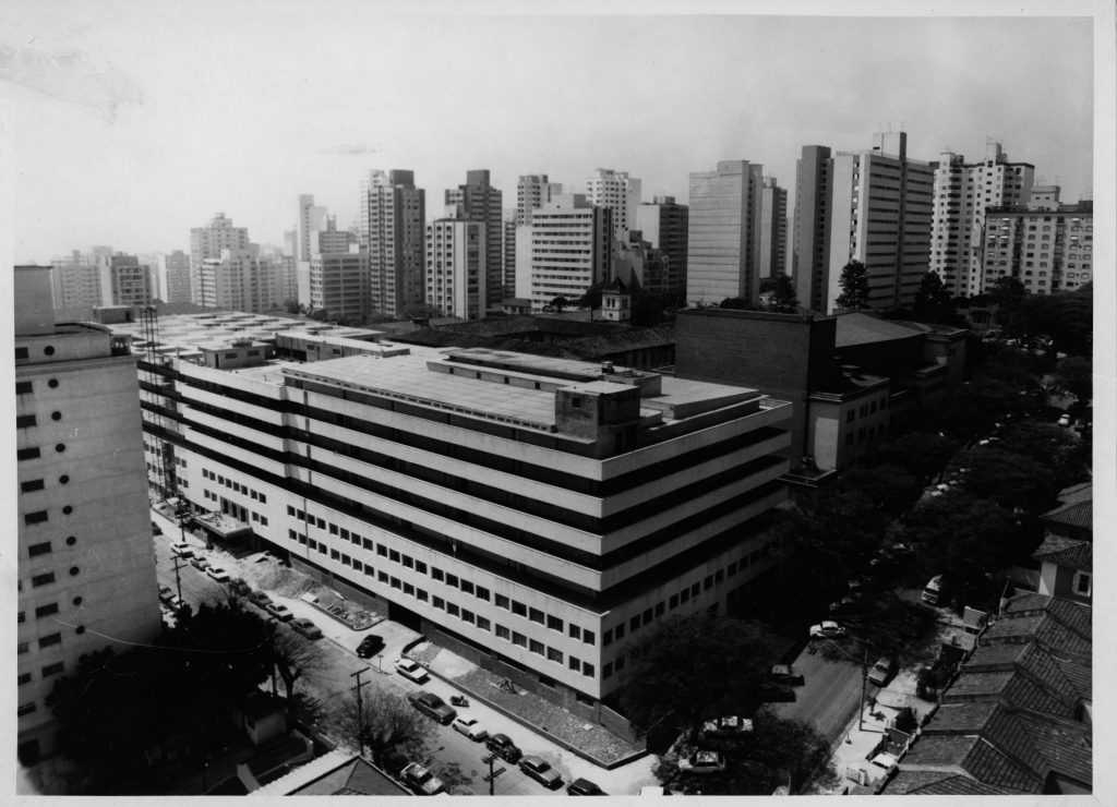 Foto em preto e branco de prédio com os dois primeiros andares com várias janelas e os 5 últimos com grandes vãos. No fundo vemos vários edifícios residenciais.