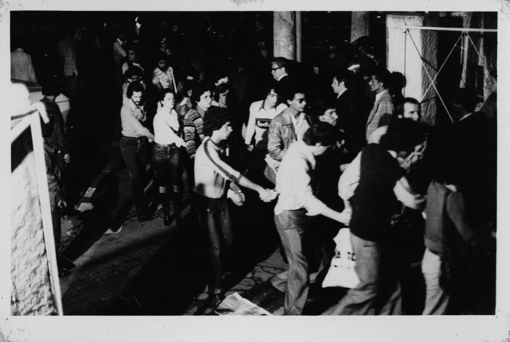 Foto em preto e branco onde vemos vários estudantes detidos em uma fila indiana e de mãos dadas saindo de um portão com alguns homens de terno observando eles passarem e serem encaminhados.