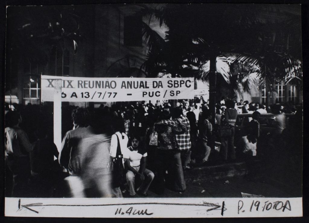 Foto em preto e branco onde vemos um cartaz com os dizeres “Reunião Anual da SBPC 6 a 13/7/1977 - PUC - SP” e várias pessoas sentadas ou caminhando perto deste cartaz e entre palmeiras em frente a um edifício.