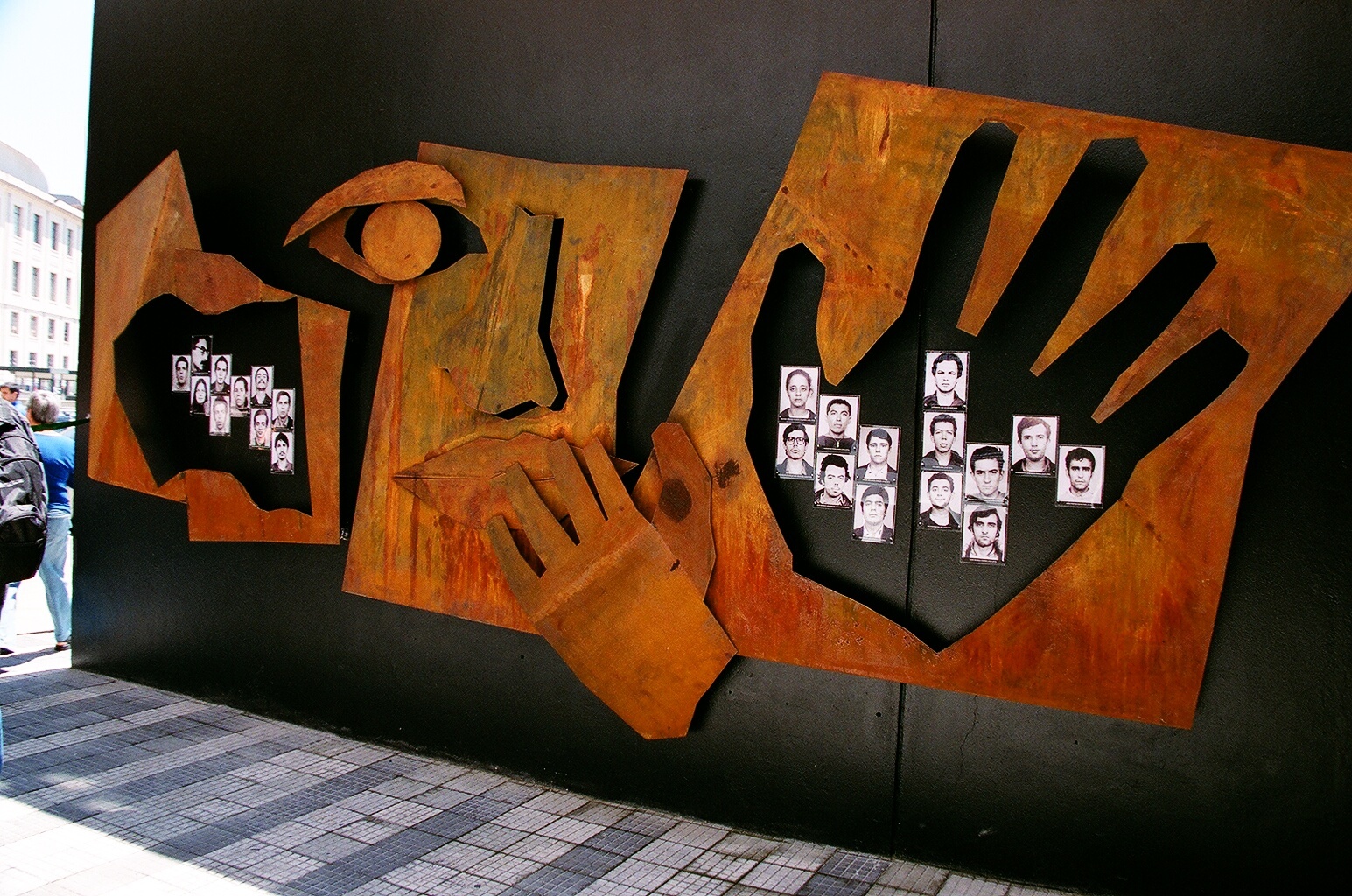 Foto colorida de instalação artística feita em ferro da cor ferrugem com a representação estilizada de um rosto e 2 mãos. Por dentro de uma das mãos, vemos 14 fotos 3x4 de jovens.
