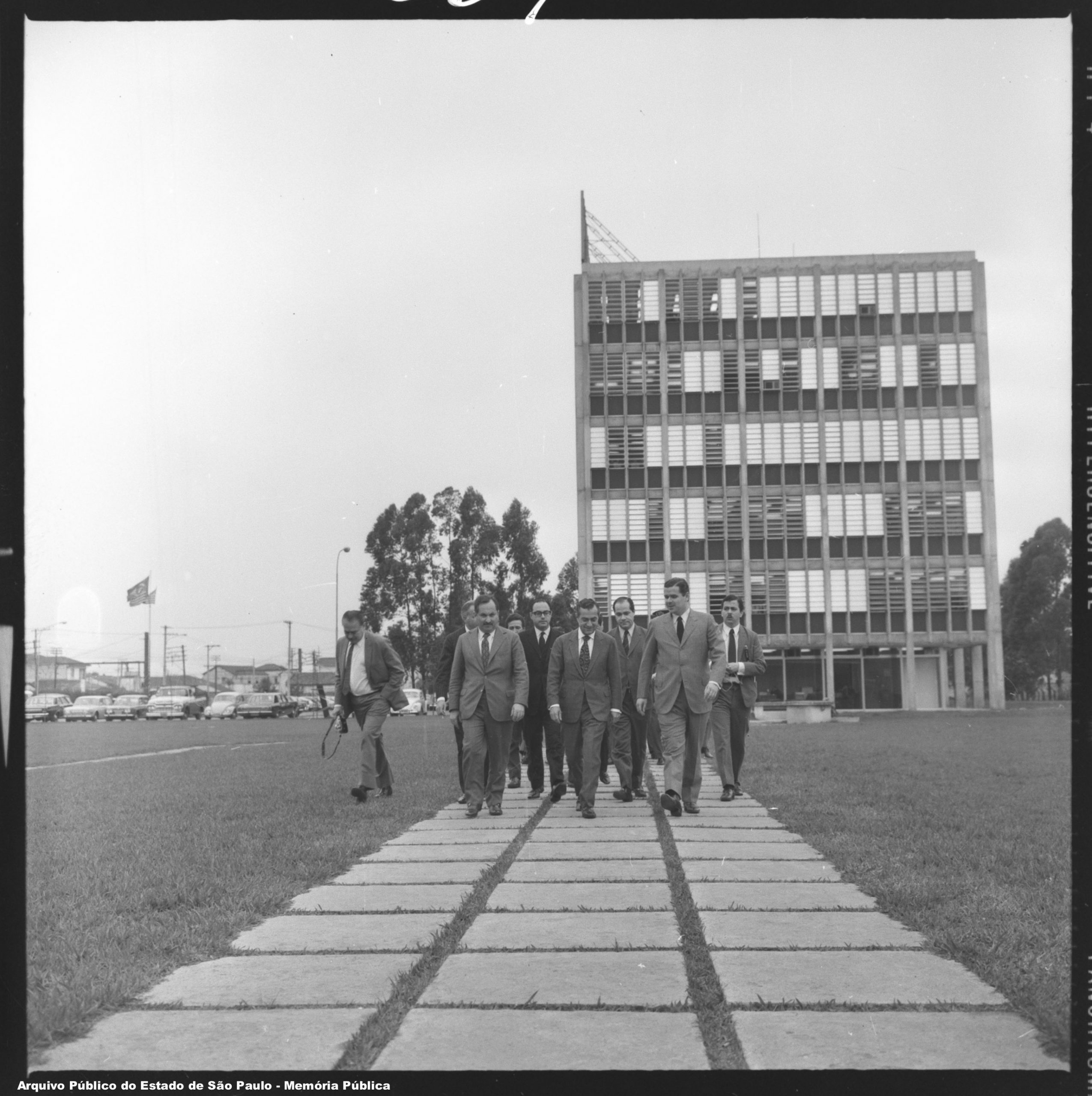 Foto em preto e branco. Dez homens de terno e gravata andam em frente por um caminho de cimento num gramado. Ao fundo há um prédio de escritório de seis