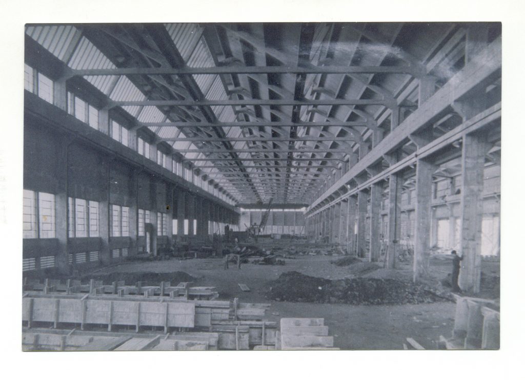 Foto em preto e branco do interior de um galpão em construção. Mostra as vigas de sustentação no teto e nas paredes, janelas do lado esquerdo, alguns homens trabalhando, um monte de terra à frente e estruturas de cimento e madeira.