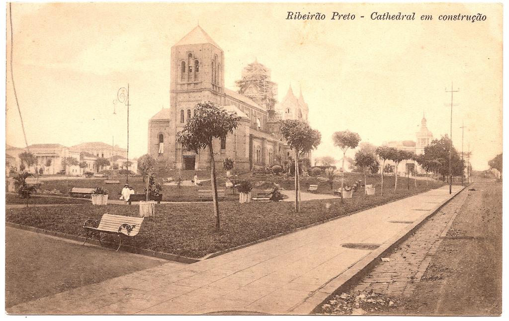 : Foto em preto e branco de uma praça com gramados e bancos. No centro da imagem vemos uma grande igreja, com a torre central ainda em construção.
