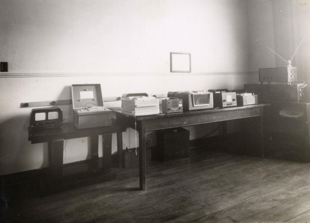 Foto em preto e branco de uma sala com tábua corrida e duas mesas encostadas na parede. Sobre as mesas vemos aparelhos eletrônicos quadrados antigos.