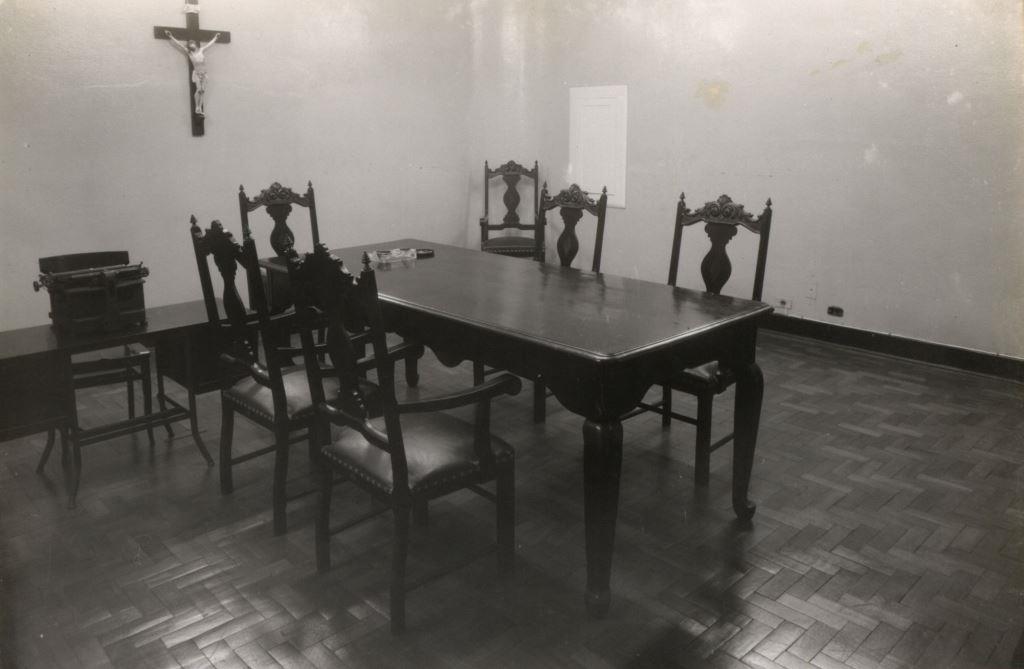 Foto em preto e branco de um salão com uma mesa de reunião retangular e 6 cadeiras entalhadas ao redor. No fundo, pregado na parede, um crucifixo grande.