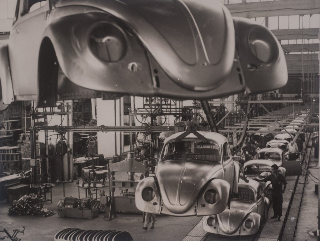 Foto em preto e branco de linha de produção de uma fábrica que apresenta a estrutura metálica de fuscas. Estas estruturas estão enfileiradas e uma máquina suspende 2 carros no primeiro plano.