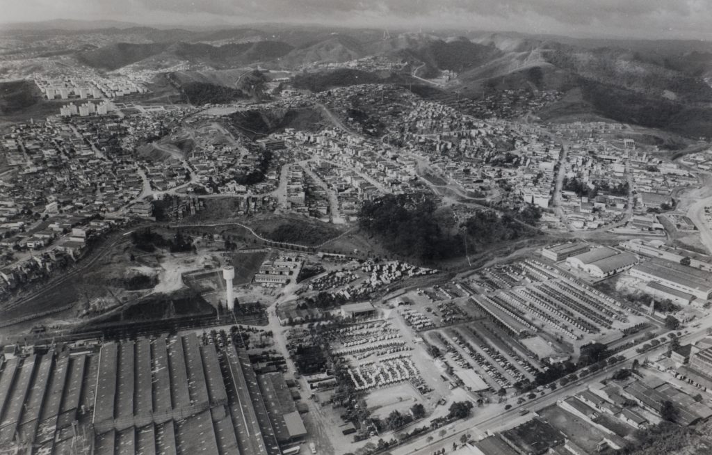 Foto aérea em preto e branco de cidades construídas entre vales e colinas. Nosso ponto de vista enxerga os telhados e as divisões das ruas. No primeiro plano vemos as coberturas de uma fábrica.